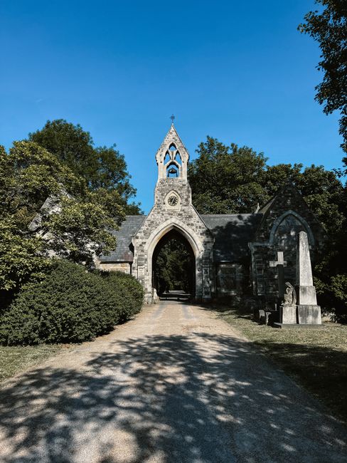 An exterior view of a historic stone church with a pointed arch entrance, clock face, and a small bell tower, surrounded by lush green trees and shrubbery. The gravel pathway leading to the church is 
