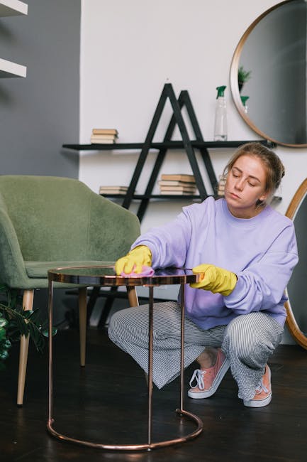 A young woman with blonde hair tied back is engaged in surface cleaning in a modern living room. She is wearing a lavender long-sleeve shirt and yellow rubber gloves, and is using a pink cloth to wipe