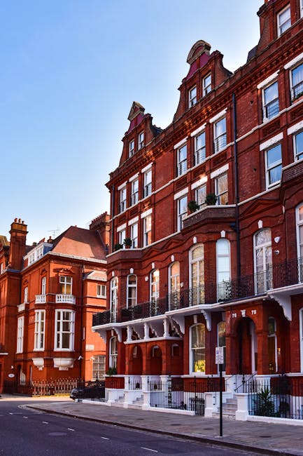 Photograph of a row of historic red brick residential buildings along a quiet street in Ealing, London, under clear blue skies. The buildings feature large sash windows, ornate white detailing around the windows and roofline, and small balconies with black wrought iron railings. The street is clean with no visible litter, and the pavement appears well-maintained. Shadows from the buildings indicate bright sunlight, enhancing the vibrant red tones of the brickwork. The scene reflects a tidy, well-preserved urban environment, suggesting ongoing maintenance and cleanliness, in line with the professional cleaning services offered by Cleaners Ealing for domestic and commercial properties in the area.