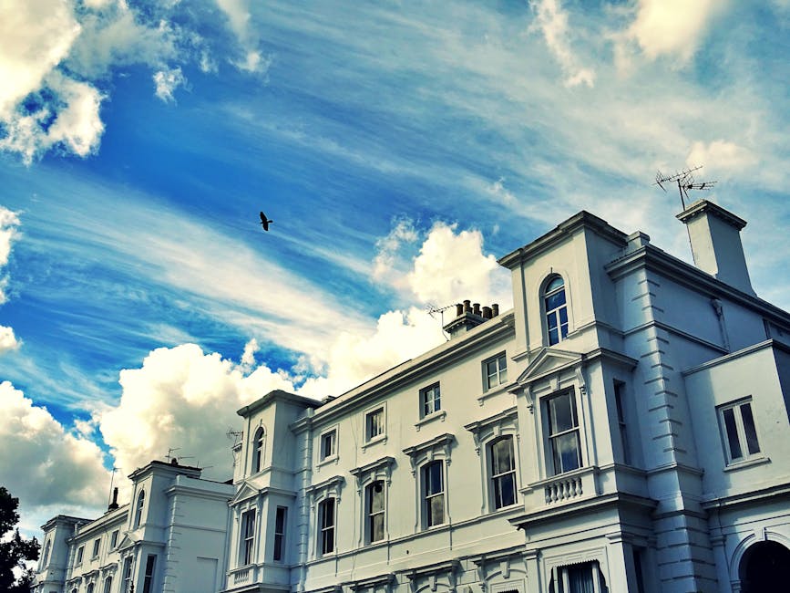 A white, multi-story residential building featuring classic architectural details such as decorative cornices, window surrounds, and a small balcony. The building's exterior walls are smooth and well-maintained, with clean, clearly visible windows that reflect the sky. The roof has chimneys and television antennas, and the scene is set against a vibrant blue sky with scattered white clouds. A bird is flying nearby, and the overall appearance suggests a tidy and well-kept property, consistent with the attention to cleanliness promoted by Cleaners Ealing, specialists in domestic and commercial surface cleaning and sanitisation services.